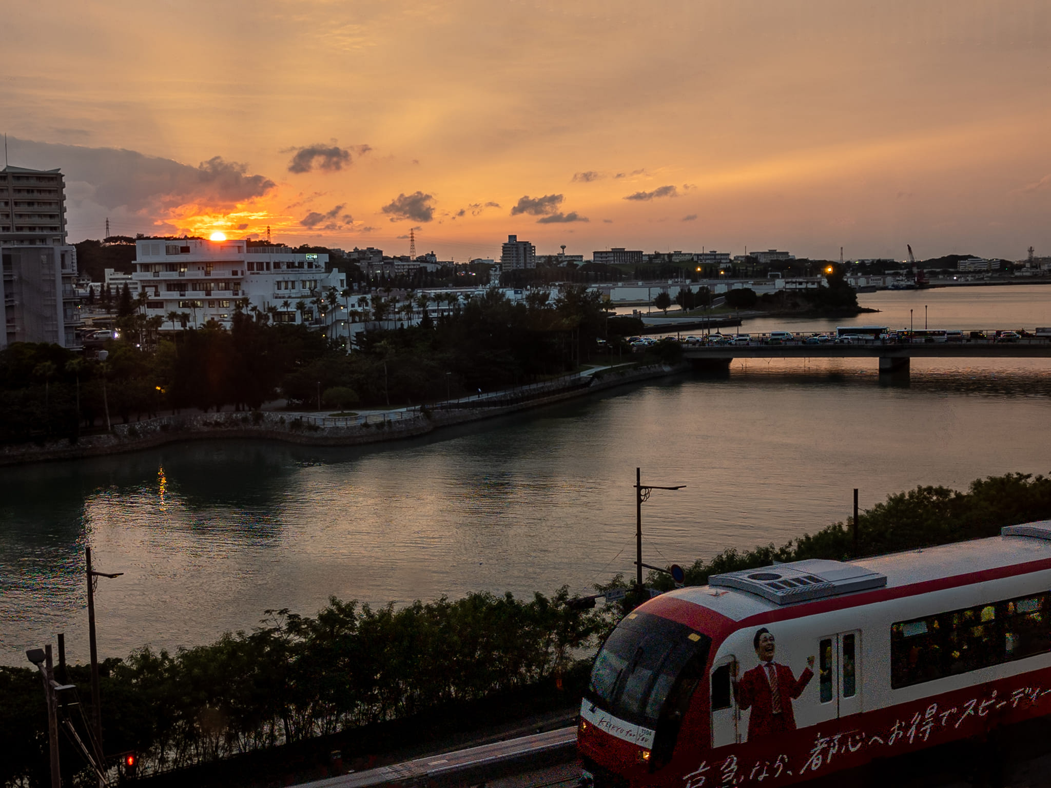 The sun sets over some naha buildings in the left of the image as a naha monorail goes by towards the bottom