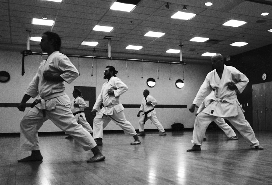 Students dressed in karate uniforms practice the first kata of Matsubayashi-shorin ryu. They are facing the left side of the photo, which is in black and white.