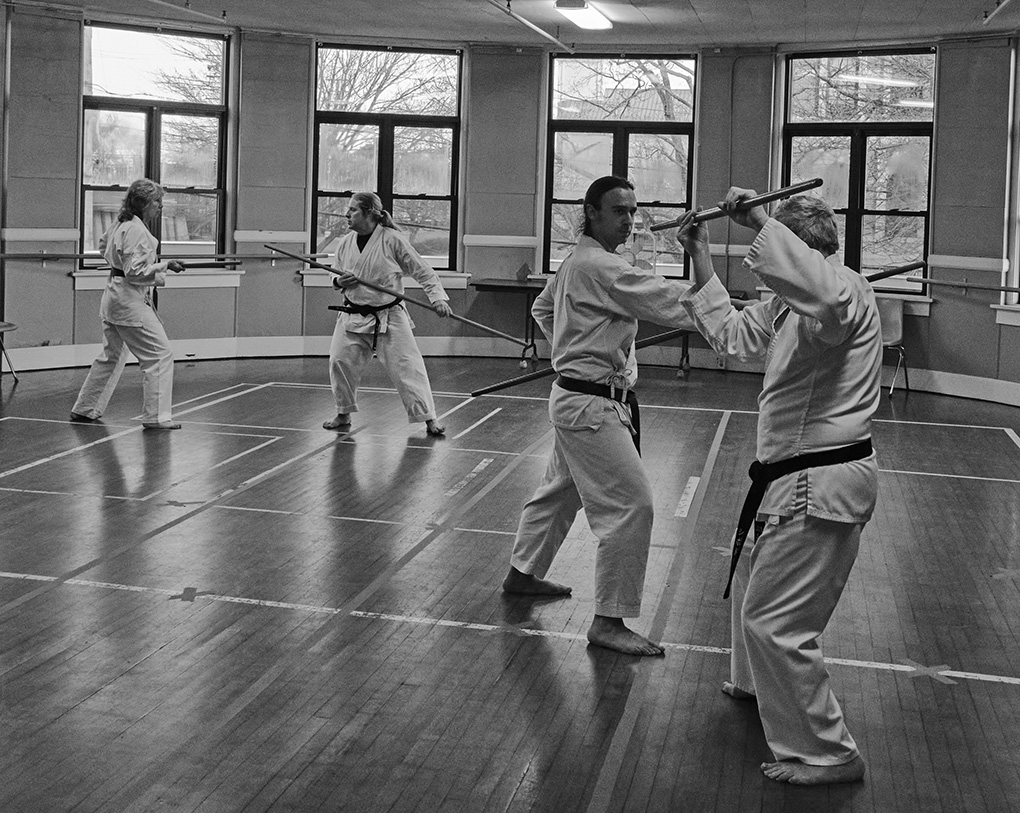 In this black and white photo, four students practice traditional kobudo. they attack and defend themselves using the bo, or 5 foot staff.
