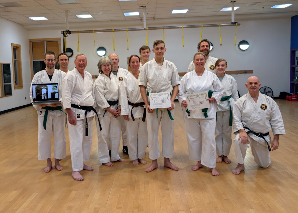 12 Karate students pose with the one on the left holding a laptop picture a 13th student, and two students in the center holding new certificates