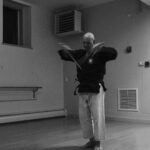 Black and white image of a karateka standing and yielding two blurry sais. His top is black and his pants are white