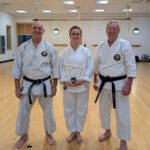 3 karate students face the camera. Two with black belts, the 3rd has receievd a certificate and green tips