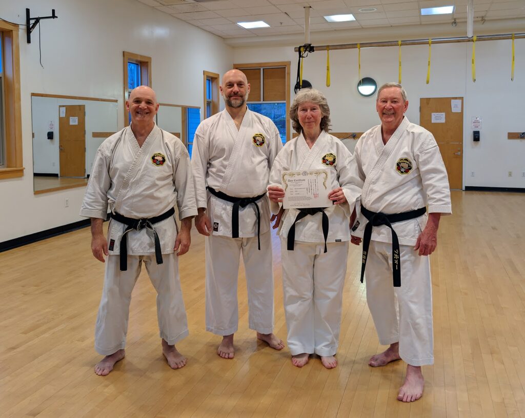 Four karate students in traditional uniforms and black belts pose for the camera. One of them has just received a certificate.