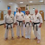 Four karate students in traditional uniforms and black belts pose for the camera. One of them has just received a certificate.