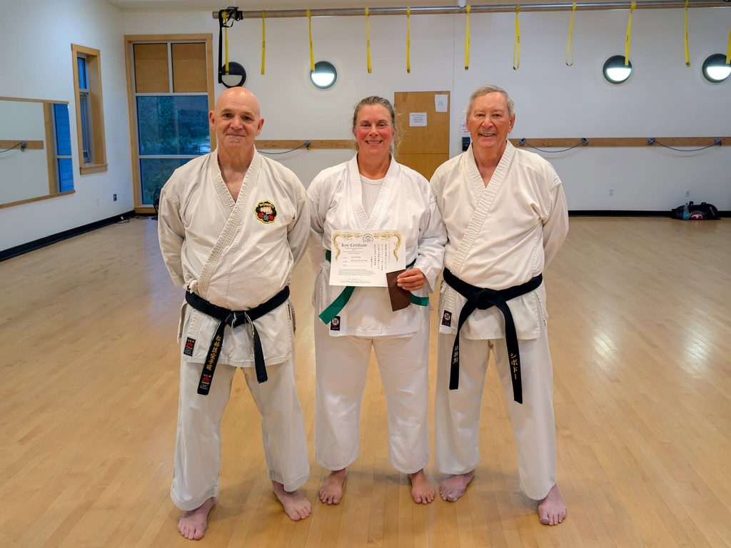 Three karate students pose for a photo. Two male black belts surround a female green belt holding her certificate for brown tips