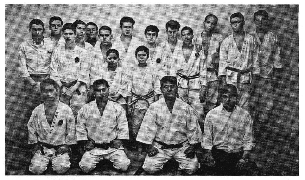 Four karateka in uniform in the front row seated with 15 karateka standing in the back